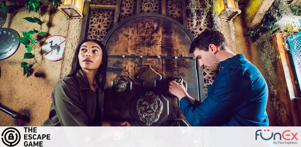 A woman and man are engaged in an escape room activity. The woman looks upwards, thinking, while the man examines a clue closely. They're surrounded by rustic decor, including a wall-mounted telephone and cobwebs. The Escape Game by FunExpress logo appears in the corner.