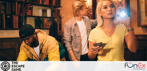 Image depicts three individuals engaged in an escape room activity. To the left, a person in a yellow jacket examines clues on a table. In the background, a participant is looking away thoughtfully. To the right, a woman in a yellow top uses a flashlight, possibly searching for hidden messages or objects. The room has a vintage feel, with books and warm lighting. The logos for The Escape Game and FunEx are visible, suggesting a collaborative experience.