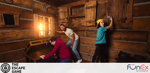 Image of three individuals in an escape room with rustic wooden walls. A person in a teal shirt is reaching up towards a high shelf, another in a white shirt is assisting, and a third in a maroon top is examining something on a wooden desk. The room is warmly lit, creating an immersive, puzzle-solving atmosphere. The logo for The Escape Game and FunEx are displayed on the lower right.