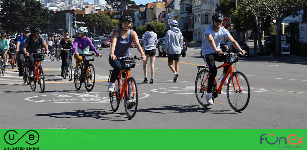 This image features a group of cyclists enjoying a sunny day on a city street. There are several people riding bicycles in the foreground, all wearing casual attire and helmets for safety. The cyclists are riding within a marked bike lane, as indicated by the painted symbols on the pavement. The background portrays a residential area with a clear view of houses and parked cars, alongside pedestrians walking on the sidewalk. The sky is clear and blue, suggesting favorable weather conditions for outdoor activities. The lower part of the image displays two logos: "UB Unlimited Biking" on the left and the FunEx.com logo on the right, identifying the image's association with FunEx and a biking service or promotion.

Experience the joy of outdoor cycling adventures and save on your next trip with FunEx – offering the lowest prices on tickets for memorable experiences!