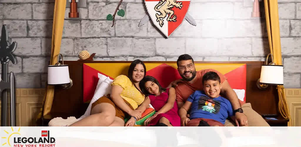 A family of four is smiling on a bed, with medieval-themed decorations in the background. Mom and dad are cozied up with two happy kids. LEGOLAND New York Resort branding is visible.