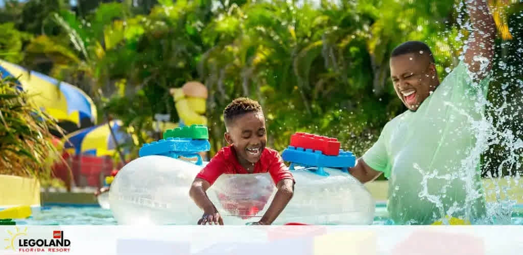 Image shows a joyful scene at a colorful Legoland Florida Resort water park. A child and an adult are playing with large Lego brick float toys in the water, both smiling with visible delight. The background is sunny with lush greenery and vibrant water slides, creating a lively atmosphere for family fun.