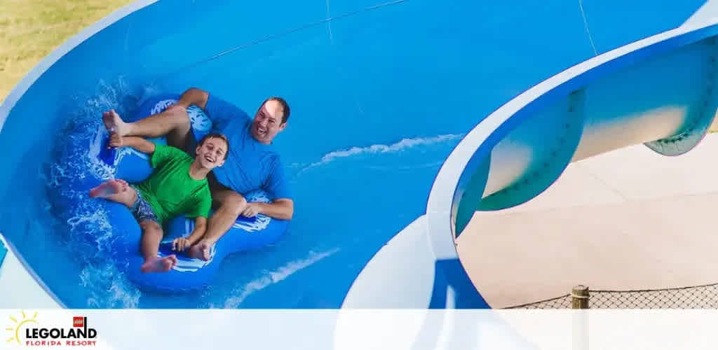 Two people are joyfully sliding down a bright blue water slide at LEGOLAND Florida Resort. They are sharing a double tube, splashing water around as they descend. The sky is clear in the backdrop, enhancing the thrilling atmosphere of the water park.