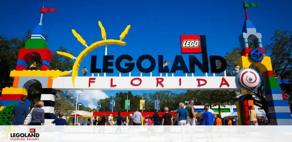 Entrance to LEGOLAND Florida Resort with a bright, multi-colored archway spelling out LEGOLAND FLORIDA in large block letters. The arch is adorned with LEGO sun and clock decorations, with visitors walking underneath on a sunny day.