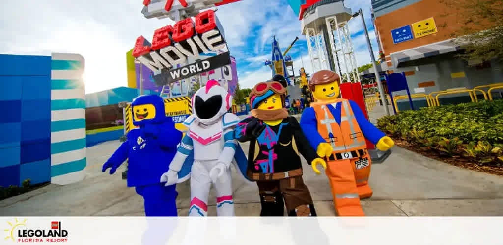 A vibrant LEGOLAND Florida Resort entrance with life-sized LEGO characters posing. From left to right, a blue astronaut, a white and pink space character, a pink and purple pirate girl, and an orange construction worker welcome guests under a colorful LEGO MOVIE WORLD sign. Bright park buildings provide a cheerful backdrop.