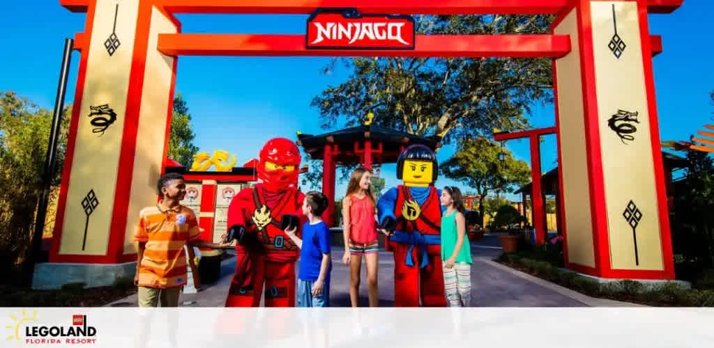 A vibrant scene at LEGOLAND Florida Resort showing visitors interacting with life-sized LEGO figures resembling Ninjago characters, set against a large red and yellow Asian-inspired gateway that reads 'NINJAGO' on top. The sky is clear and the mood is cheerful.