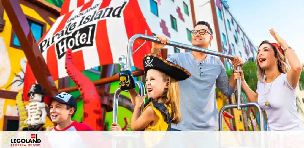 A joyful family of four is engaging in pirate-themed fun outside the colorful LEGOLAND Pirate Island Hotel. A young child dressed as a pirate and an excited boy in a baseball cap accompany smiling adults on a bright sunny day.