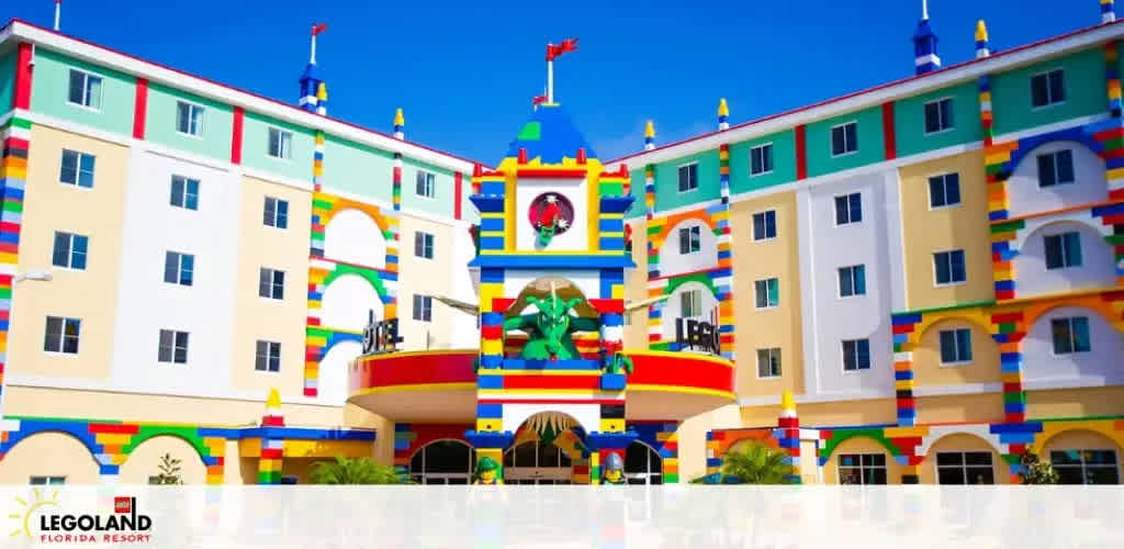 Image of a vibrant, colorful facade of a Legoland Florida Resort hotel designed to mimic the appearance of Lego bricks. The building features a whimsical clock tower and playful Lego figures decorating the exterior. Clear skies and the resort’s branding are visible.