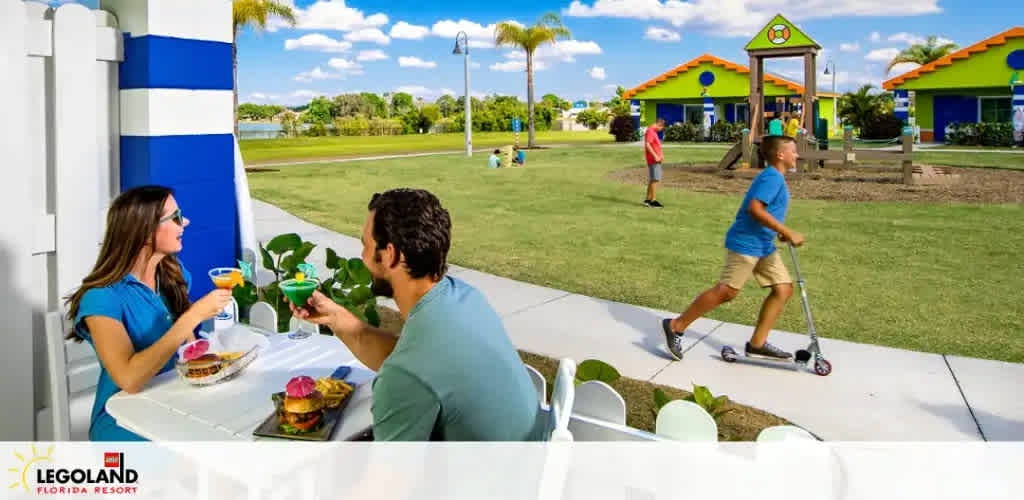 Image shows a sunny outdoor scene at LEGOLAND Florida Resort. In the foreground, a couple enjoys beverages at a table, while behind them, a child rides a scooter. Colorful buildings with green roofs create a vibrant background with clear blue skies above.