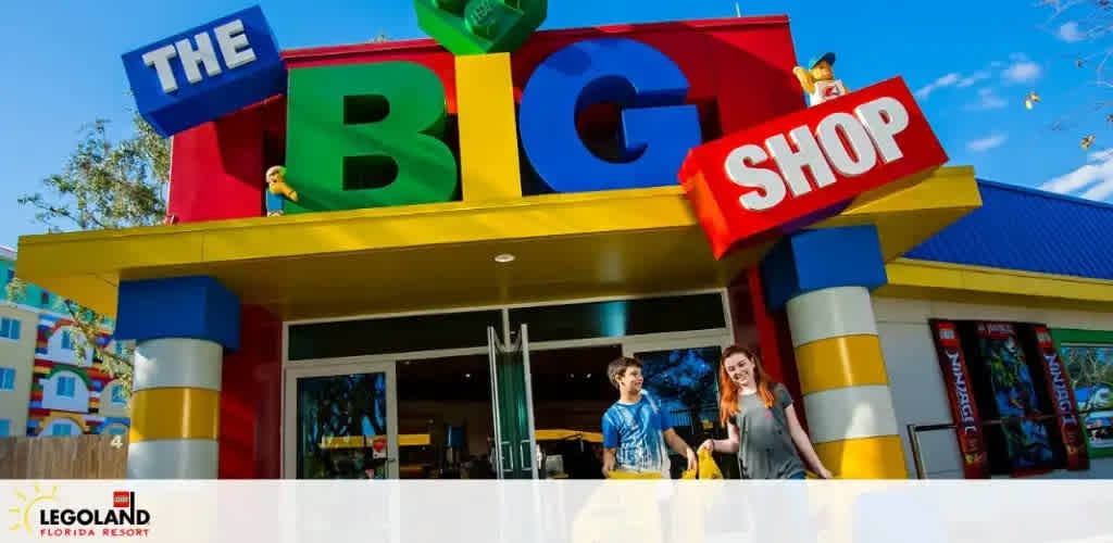 Image shows the entrance of The Big Shop at LEGOLAND Florida Resort. Oversized colorful LEGO-like blocks spell out the sign above the doorway. Two individuals exit, smiling. Bright blue skies and a playful, vibrant aesthetic set a cheerful tone for the family-friendly shopping experience.