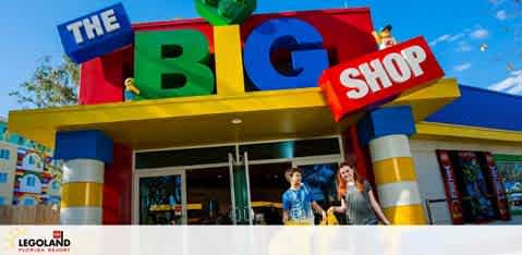 Image shows the entrance to The Big Shop at LEGOLAND, featuring oversized LEGO blocks spelling the shop's name. Bright colors adorn the facade while two visitors are exiting, with toys visible inside. Clear skies in the background.