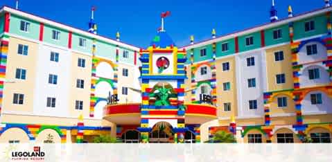 The image shows a vibrant, multi-story hotel facade with a playful, colorful Lego theme. The architecture includes bright primary colors, with large Lego brick designs adorning the entrance and surrounding the windows. The central portion is marked by a large red Lego heart framed by green and yellow bricks. The sky is clear and blue, complementing the cheerful atmosphere of the building. The LEGOLAND logo is visible at the bottom left, indicating the hotel's association with the amusement brand.