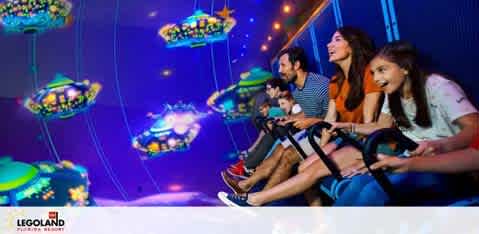 A dynamic scene at LEGOLAND Resort featuring four excited guests on a ride. The riders, two adults and two children, are seated in a roller-coaster-like cart, secured by safety bars. They're immersed in a vibrant space-themed environment, with colorful, illuminated LEGO spacecraft above. Their expressions and body language exude enjoyment and thrill.