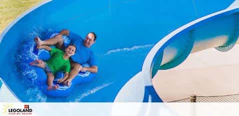 Image showing two individuals, an adult and a child, joyfully sliding down a curvy blue water slide at Legoland Florida Resort. They are sitting on a double raft, with the adult at the back. Splashing water and their delighted expressions convey a sense of fun and excitement.