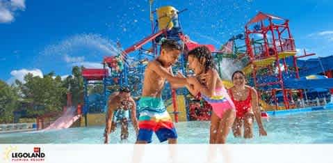 Children play in a vibrant water park under sunny skies. In the foreground, two kids engage in a playful splash fight in shallow water. Behind them, others enjoy the splashing structures and slides. The image evokes a joyful atmosphere at a Legoland Resort.