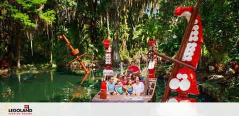 Family and friends enjoy a boat ride at LEGOLAND Florida, surrounded by lush greenery with LEGO sculptures, such as a friendly red and white turret with a smiling face, enhancing the playful atmosphere of this aquatic adventure.