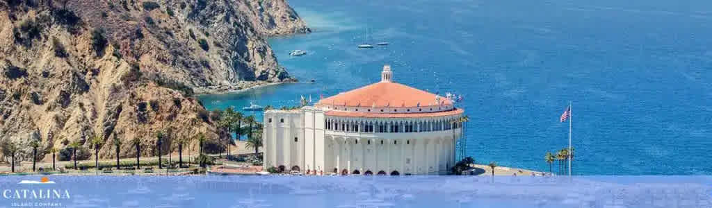 The image showcases the iconic Casino Building on Catalina Island, featuring its distinctive round structure with a red-tile roof. Surrounding the building are palm trees, azure waters, and anchored boats. The American flag flies proudly at the waterfront. The word 'CATALINA' is overlaid at the bottom left.