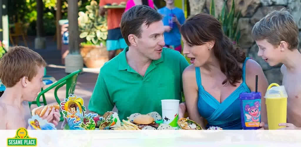 A family enjoying a meal at Sesame Place. Two children on the left, one eating a hotdog, and adults in the middle, smiling and conversing. One child on the right looks at the woman, holding a drink. Theme park dining area with foliage in the background.