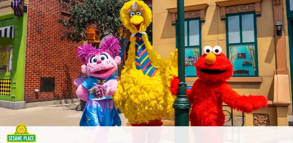 Image shows three colorful Sesame Street characters in a sunny outdoor setting, likely a theme park. From left to right: a pink character with a tuft of colorful hair and wearing a blue dress, a tall yellow character sporting a striped tie, and a smiling red character. They stand in front of a façade resembling city buildings. The logo at the bottom indicates the location as Sesame Place.