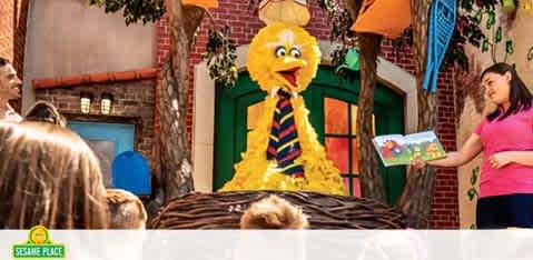 Image depicts a scene at Sesame Place with a large, cheerful yellow bird character greeting guests. A staff member in pink is holding a book, engaging with the visitors, who include children and adults. The backdrop features colorful, themed buildings. The atmosphere is lively and family-friendly.