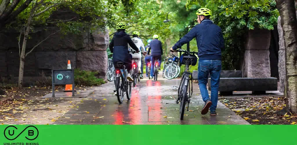 Three cyclists on a bike path lined with trees and benches, with a wet surface and fallen leaves.