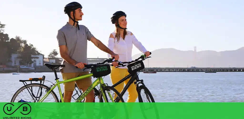 Two people with bikes by the water, under a clear sky, with hills in the distance.