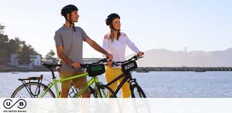 This image features a man and a woman standing with bicycles by a serene waterfront during what appears to be late afternoon. The man is on the left, dressed in a casual grey t-shirt, black shorts and wearing a helmet and sunglasses. He is holding a dark green bicycle. The woman is on the right, wearing a white blouse, beige shorts, a helmet and also has sunglasses on. She holds a yellow bicycle. Both are looking to the side with a view of water in the background and a faint outline of hills in the distance. There's a warm glow suggesting sunset or sunrise, contributing to the tranquil atmosphere. The bicycles have bags on the front handlebars indicating preparedness for a day of adventure or travel.

At FunEx.com, we pride ourselves on offering the best deals and lowest prices on tickets to a wide variety of exciting activities, ensuring that your next adventure is not only memorable but also comes with great savings.