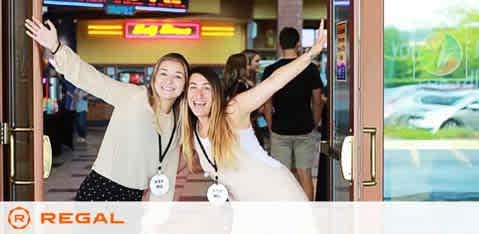 Two joyful individuals are exiting a Regal movie theater, their arms raised high in a welcoming gesture. They're wearing badges and smiling brightly, indicating they may be theater staff or part of a promotional event. The entrance showcases the theater's branding, with a digital display visible in the background. The parking area outside is partly seen through the doors.