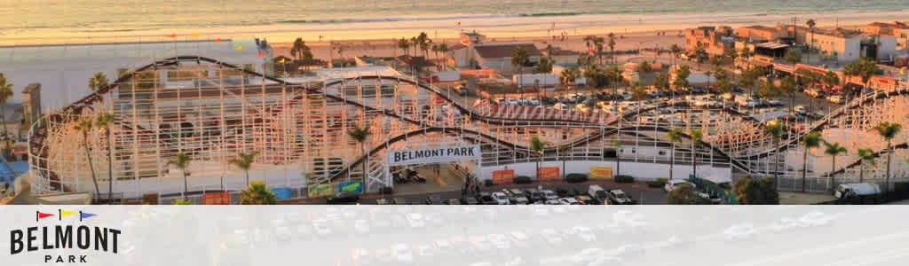 Image shows an expansive wooden roller coaster at Belmont Park amusement park near the beach during sunset. The coaster structure with its twists and turns dominates the foreground, while the beach and ocean are visible in the background. The sky is tinged with warm hues of the setting sun. The Belmont Park logo is visible on the lower left side.