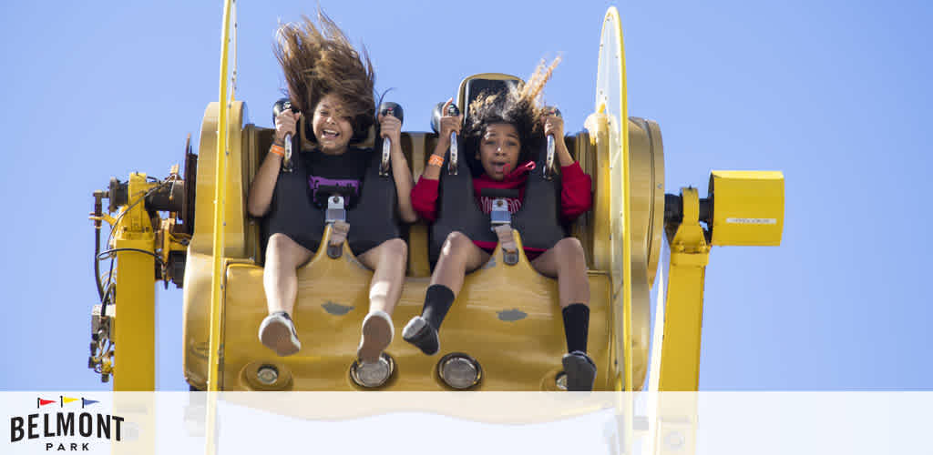 Two thrilled individuals ride a yellow rollercoaster against a clear blue sky. Their hair and expressions capture the excitement of the descent. Below, the logo of Belmont Park suggests the location of this fun experience.