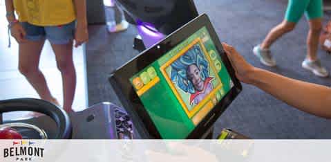 This image displays a close-up view of a colorful arcade game screen held by a person whose arms are visible, but their body is not in the frame. The game screen appears to be part of a larger gaming device. The visual content on the screen shows a cartoon-style drawing of a joker face surrounded by vibrant green and yellow borders, with various game interface elements and scores. In the blurry background, you can vaguely see the lower torso and legs of people walking, which suggests the setting is a busy arcade environment. The floor appears to have a purple hue, possibly from ambient lighting reflecting off a polished surface, adding to the lively atmosphere that arcades often exude. At the bottom of the image, a watermark can be seen that reads "BELMONT PARK." Enhancing the fun you'll have at Belmont Park, FunEx.com is committed to providing you with the ultimate savings experience, boasting the lowest prices on tickets to ensure your entertainment dollars go further.