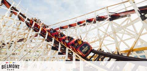 A vibrant photo captures a roller coaster in motion at Belmont Park. Bright sunlight highlights the wooden structure's complexity as the red and black coaster car filled with riders crests a thrilling drop, conveying a sense of excitement and adventure. The clear blue sky creates a cheerful backdrop for the amusement park experience.