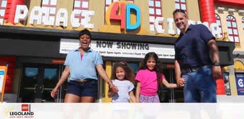 A happy family consisting of two adults and two children stand in front of the 4D theater at LEGOLAND. The theater boasts large letters on the facade and a marquee announcing 'NOW SHOWING'. The adults are on either side of the grinning children. The building resembles a colorful Lego structure, adding to the playful atmosphere of the scene.