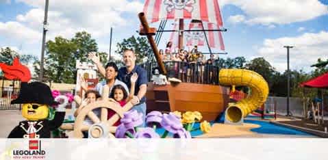 The image features a joyful family on a bright day at LEGOLAND. A man and two children, one steering, play on a large pirate ship playground with vibrant LEGO figures and a slide. A smiling mascot stands on the left. The atmosphere is cheerful and playful.