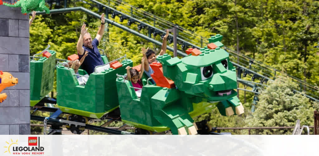 Image features visitors enjoying a ride on a green dragon-themed roller coaster at a LEGO-themed park. The front of the coaster resembles a dragon's head with playful eyes and teeth, crafted to look entirely made from LEGO bricks. Trees and vegetation surround the track, enhancing the outdoor adventure atmosphere. Excitement is evident on the faces of riders with arms raised in joy.