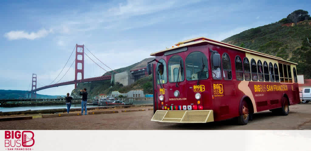 This image features a panoramic view of a scenic location, with a prominent red sightseeing trolley bus parked in the foreground on the left side of the image. The words "BIG BUS SAN FRANCISCO" are visible on the side of the vehicle, which has multiple open windows for an unobstructed touring experience. In the background, the iconic Golden Gate Bridge stretches across the scene, its red suspension towers rising high above the horizon under a partly cloudy sky. Two individuals are standing to the right of the bus, near the waterfront, taking photos of the picturesque setting. Gentle hills and a serene bay complete the landscape, offering a blend of natural and man-made marvels.

Remember to check out FunEx.com for exclusive savings and the lowest prices on tickets for your next adventure with Big Bus San Francisco.