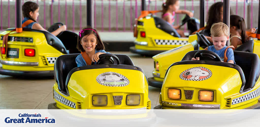 This image displays an amusement park ride from California's Great America featuring children enjoying mini yellow bumper cars. A smiling girl with dark hair wearing a safety belt appears in the foreground, holding onto the steering wheel of her car with a look of joy and anticipation. Behind her, other children are seated in similar cars, some waiting for the ride to start while others are already navigating their vehicles. The cars have fronts designed to mimic classic yellow taxis, including checkered details and headlight graphics. Each car has an image of a cartoon character resembling a dog with a police hat on the front and back. The environment is a covered area with a burgundy and white striped awning, and the floor is a smooth, grey surface suitable for the cars to glide over.

FunEx.com offers exclusive discounts, ensuring you enjoy the lowest prices on tickets for family-friendly destinations like California's Great America.