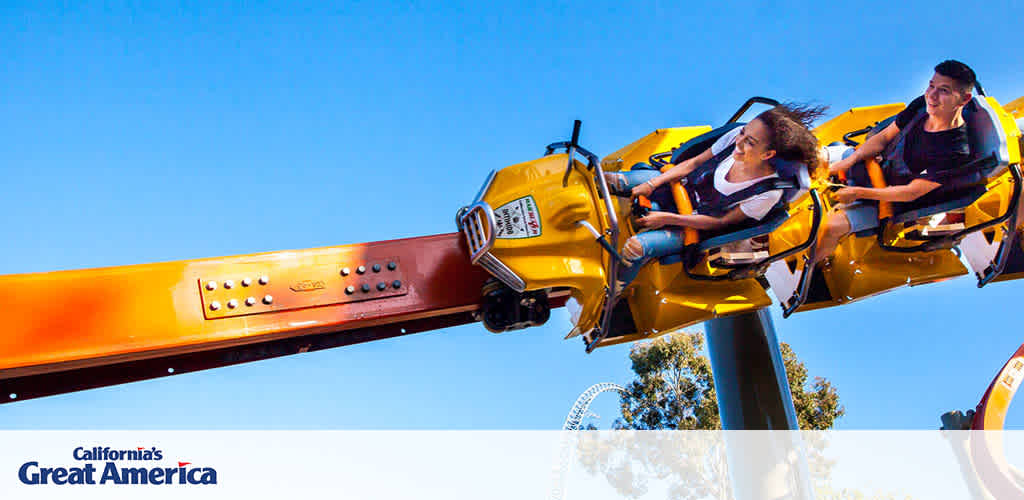 This image exhibits a clear blue sky background with an amusement ride from California's Great America in the forefront. The ride, featuring a brightly colored yellow and orange mechanical arm, is in motion and has two delighted riders secured in an open, roller-coaster style car. The riders appear to be experiencing the thrill of the ride, with their hair flowing backward due to the movement, and expressions of enjoyment visible on their faces. Lush green trees are subtly visible in the lower background, suggesting the ride is surrounded by a scenic landscape.

At FunEx.com, embark on an adventure of thrills and excitement while enjoying the savings that come with our lowest prices on tickets - because every moment should be as enjoyable as the attractions themselves.