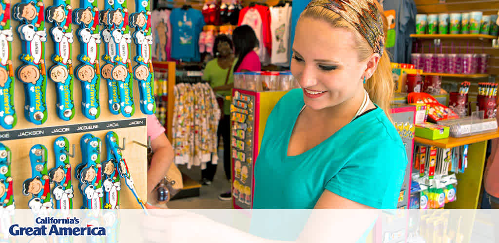 This image features a smiling woman standing in a brightly colored gift shop at California's Great America. She is interacting with a display of personalized items, in particular focusing on keychains with various names printed on them. The keychains are adorned with images of cartoon characters and hang on a wooden display board. The background includes an assortment of merchandise such as clothing, toys, and drinkware neatly arranged on shelves. In the corner, the logo of California's Great America is visible.

Experience the excitement of California's Great America and enjoy the added bonus of savings when you purchase your tickets through FunEx.com, where we strive to offer the lowest prices!