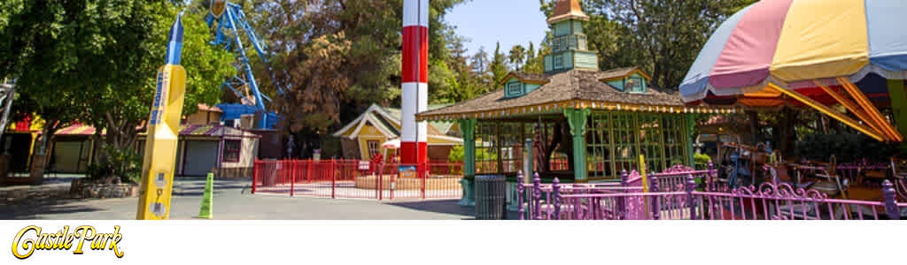 This image displays a vibrant amusement park setting, showcasing various attractions under a clear blue sky. On the left, a tall, slender attraction with blue and yellow hues reaches toward the sky, adjacent to an amusement ride with red and white striped accents. The central part of the image reveals park walkways lined by colorful storefronts with details suggesting a quaint village atmosphere. On the right side, a carousel with a green roof is partially visible under a large, multicolored umbrella canopy. The scene is framed by lush green trees, and the pavement in the foreground is wide and appears well-maintained, suggesting accessibility. The bottom left corner features text that reads "Castle Park".

At FunEx.com, our visitors always find amazing discounts, ensuring you enjoy the lowest prices on tickets to premier attractions like the one pictured.