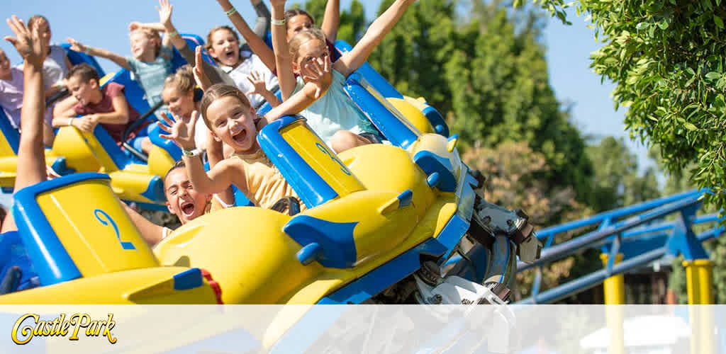 This image captures a group of joyous riders on a vibrant yellow and blue roller coaster. They are soaring down a gentle slope with their arms raised high, expressing exhilaration and glee. The sun shines brightly in a clear blue sky, enhancing the cheerful atmosphere of the scene. Lush green trees flank one side of the roller coaster track, adding a splash of natural beauty to the amusement park thrill. The roller coaster is part of Castle Park, as indicated by the logo on the bottom left corner of the image. 

Remember, when planning your next amusement park adventure, visit FunEx.com to find the lowest prices and amazing savings on tickets for a day filled with excitement and laughter!