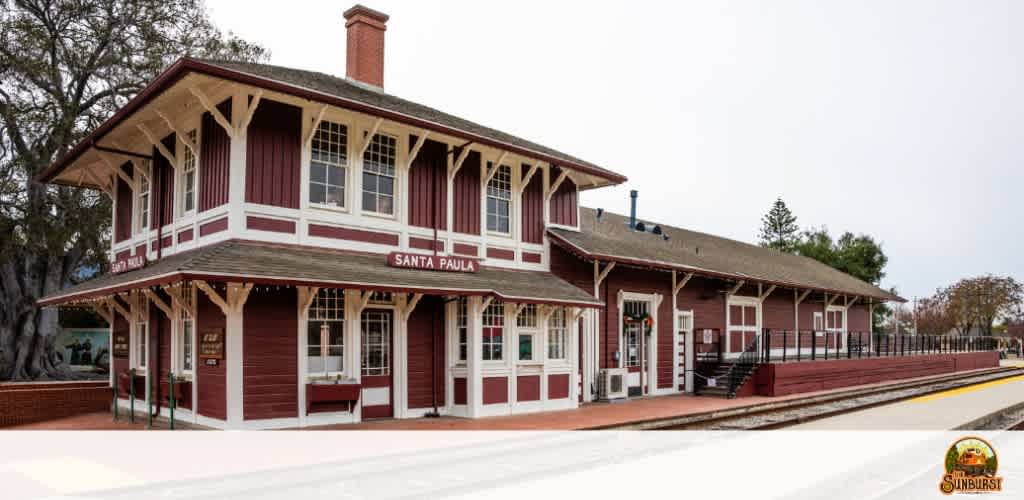 This image displays the historic Santa Paula train station under an overcast sky. This charming structure features a two-story building with contrasting white and deep red hues. The architecture includes a detailed facade with multiple windows and decorative trim. A notable chimney extends from the peaked roof, which is adorned with supporting struts adding to the building's vintage aesthetic. In the foreground, a well-maintained train platform runs parallel to the tracks, indicating the station's purpose. There is no train present in the image, and the platform appears vacant, adding to the calm and serene atmosphere of the scene. To the left, several trees can be seen in the background, suggesting a rural or small-town location. The image also includes the logos for Santa Paula and Sunburst. 

At FunEx.com, we pride ourselves on offering not just memorable experiences but also the chance for great savings - get your tickets here to enjoy the lowest prices available.