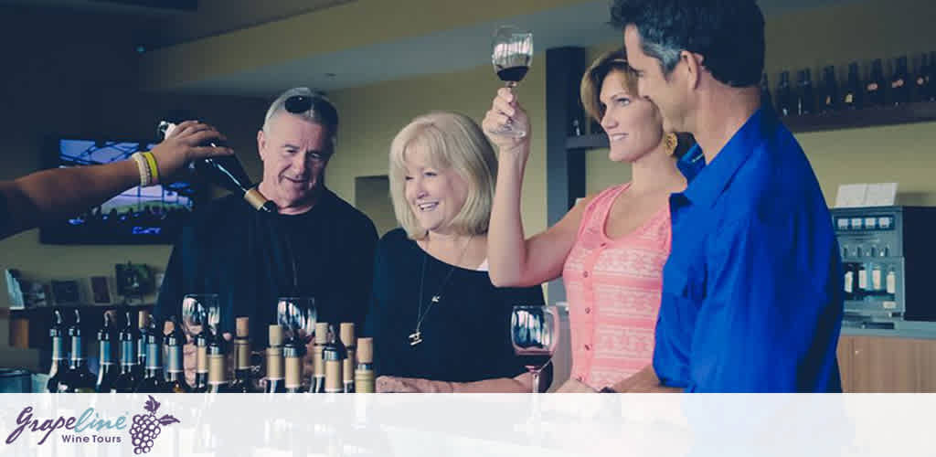 This image features a pleasant scene at what appears to be a wine tasting event hosted by GrapeLine Wine Tours. Two couples are standing at a bar, each holding a glass of red wine, with smiles on their faces, suggesting a convivial atmosphere. The individual on the far left is in the process of receiving a pour of wine from an unseen server whose arm enters the frame from the left side. The room has a modern aesthetic, with a flat-screen TV visible in the background, and a selection of wine bottles lined up in front of the couples on the bar. The GrapeLine logo is prominently displayed in the lower left corner of the image.

For our FunEx.com guests looking for an exceptional experience without straining their wallets, we're thrilled to offer incredible savings on tickets to events like these – always striving to give you the lowest prices available.