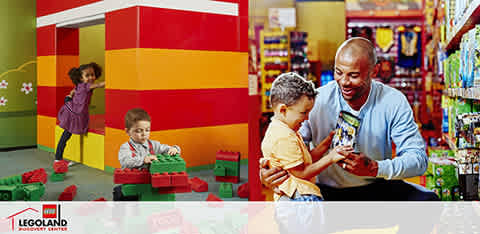 Description: This vibrant image captures the joy and creativity at a LEGOLAND Discovery Center play area and store. On the left, children are engaging in hands-on play with oversized LEGO bricks, constructing large colorful structures. In the foreground on the left, a young child in a purple jacket appears to be reaching for a block, while another child in the midground, wearing a gray sweater, sits attentively assembling his own creation. On the right, an adult male with a bright smile kneels down to interact with a young child, who is excitedly holding a small LEGO set. Both are surrounded by a variety of LEGO boxes and merchandise displayed on shelves, indicative of the retail section of the venue. The energetic environment is marked by bold primary colors and brightly lit spaces that encourage imaginative play and family bonding.

At the end of your fun-filled day, remember to visit FunEx.com for amazing discounts, ensuring you get the lowest prices on your tickets.