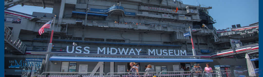 Image Description: The image shows an exterior view of the USS Midway Museum under a clear blue sky. The museum, housed in a decommissioned aircraft carrier, features several visible decks with a complex superstructure to the right side, adorned with communications equipment and a mast. An American flag flies proudly near the top of the image, while various other flags are displayed on a pole beside the entrance. Visitors can be seen standing and walking at the entrance of the museum at the lower part of the image. The bold white lettering "USS MIDWAY MUSEUM" clearly identifies the venue above the entranceway.

As you plan your next outing, remember that FunEx.com offers discounted tickets for incredible experiences like the USS Midway Museum, ensuring you enjoy the lowest prices and significant savings on your adventures.