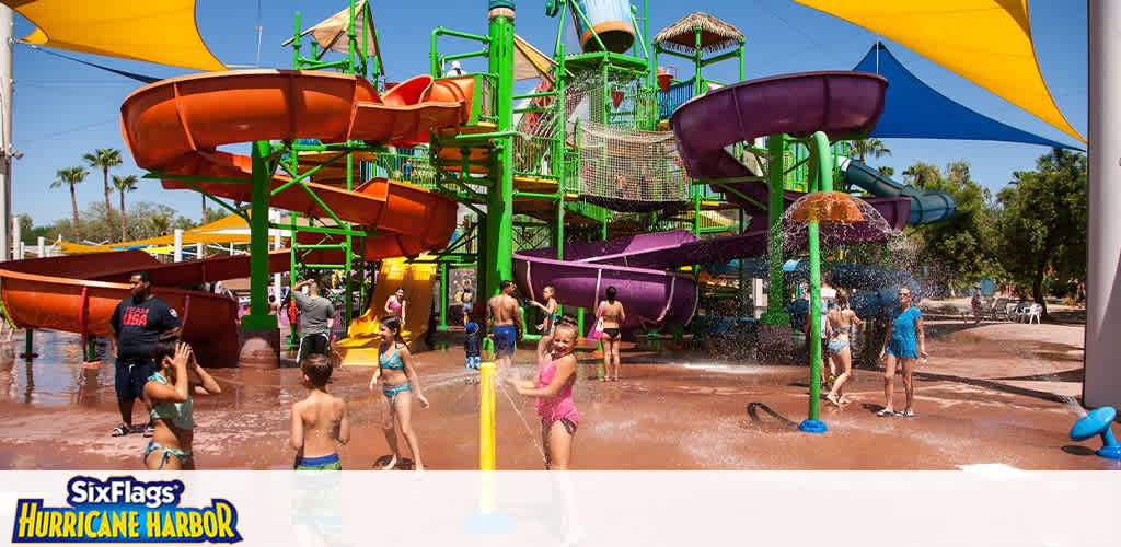 Image of a vibrant water park on a sunny day featuring a colorful array of slides in orange, green, and purple. Visitors, including children and adults, are enjoying the water attractions, relaxing under blue and yellow sunshades, and walking on wet ground. The Six Flags Hurricane Harbor logo is visible, indicating the location.