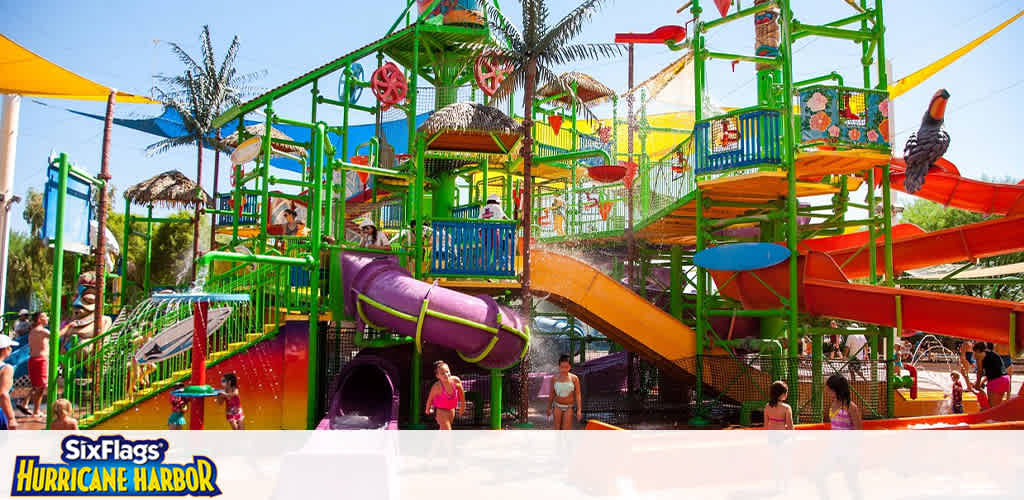 Image depicts a vibrant water playground at Six Flags Hurricane Harbor. Features include bright green, yellow, and red slides and multiple levels of play areas with sprayers. Visitors, including children and adults, are enjoying the sunny atmosphere surrounded by palm trees.