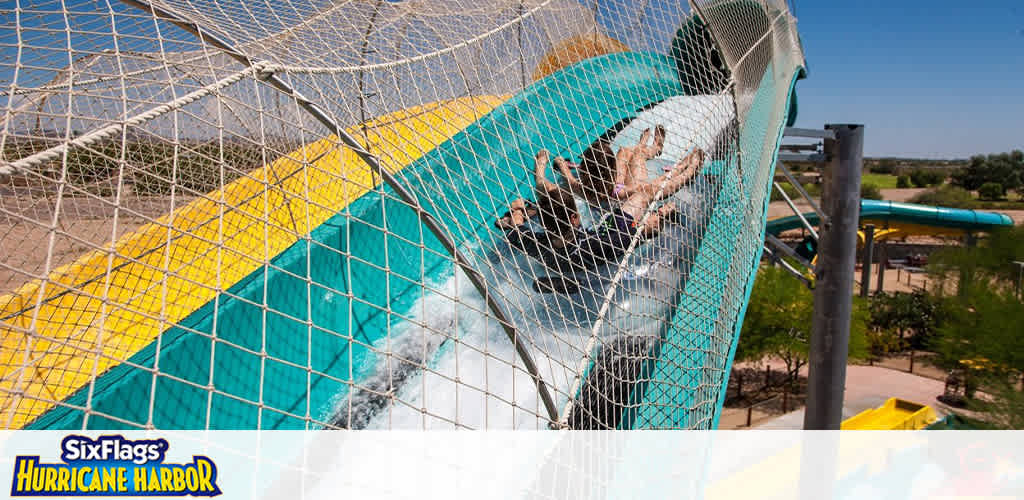Image shows a water slide at Six Flags Hurricane Harbor. People are sliding down a tube that spirals with blue and yellow stripes. Splashes of water follow their descent. Safety nets surround the slide, and the park's surroundings include greenery under a clear, blue sky.