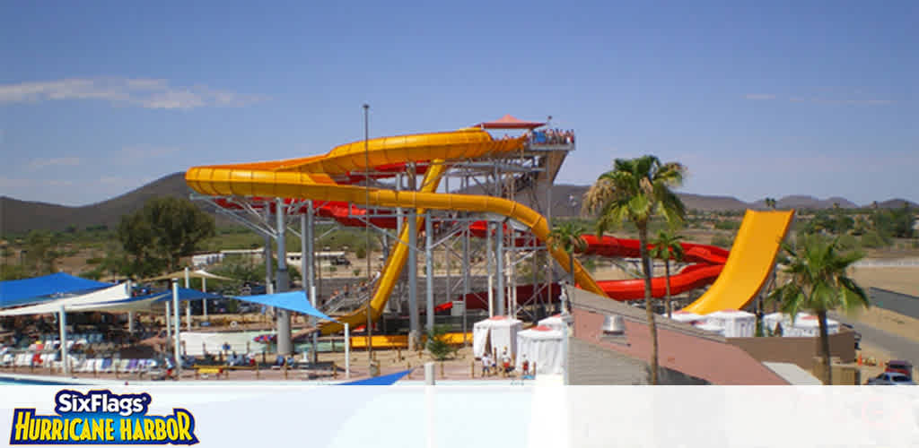 Image shows a brightly lit outdoor waterpark under a clear blue sky. At the center are tall, winding water slides in yellow and orange, towering above a pool area. People can be seen enjoying the amenities, with sun loungers and umbrellas dotted around. Lush greenery and hills are visible in the background, emphasizing the park's scenic setting. The words  Six Flags Hurricane Harbor  are overlayed at the bottom center of the image.