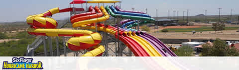 Image shows a series of colorful water slides at Six Flags Hurricane Harbor. The slides, in red, yellow, purple, and blue, twist and turn before descending downwards. The backdrop features a clear sky with faint clouds above a serene landscape with greenery and roads. The park logo is present in the corner.